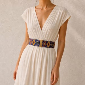 Puede incluir: Un vestido blanco con cuello en V y mangas cortas se ci&ntilde;e a la cintura con un cintur&oacute;n colorido. El cintur&oacute;n presenta un patr&oacute;n geom&eacute;trico en azul, amarillo y rojo, y una hebilla dorada.