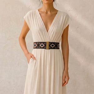 Puede incluir: Un vestido color crema con cuello en V y mangas cortas. El vestido se ci&ntilde;e a la cintura con un cintur&oacute;n negro con un estampado geom&eacute;trico en rojo, blanco y negro. El cintur&oacute;n tiene una hebilla rectangular dorada.