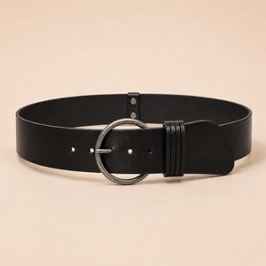 Puede incluir: Un cintur&oacute;n negro ancho con una hebilla circular de metal plateado con acabado envejecido. El cintur&oacute;n incluye una trabilla de cuero negro a juego con tres ranuras horizontales y dos peque&ntilde;os remaches cerca del cierre.