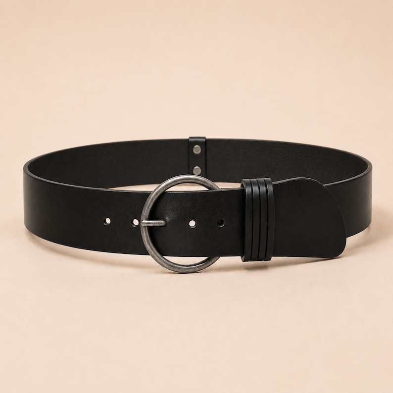 Puede incluir: Un cintur&oacute;n negro ancho con una hebilla circular de metal plateado con acabado envejecido. El cintur&oacute;n incluye una trabilla de cuero negro a juego con tres ranuras horizontales y dos peque&ntilde;os remaches cerca del cierre.