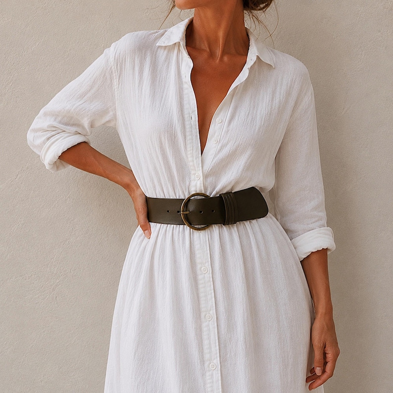 Puede incluir: Un vestido camisero blanco con botones, mangas largas y un acabado de tela texturizada. El vestido se ajusta a la cintura con un cintur&oacute;n ancho de color verde oliva oscuro que presenta una hebilla met&aacute;lica grande y redonda en tono bronce.