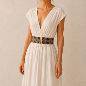 Puede incluir: Un vestido blanco con cuello en V y mangas cortas, ce&ntilde;ido a la cintura con un cintur&oacute;n estampado colorido. El cintur&oacute;n presenta un dise&ntilde;o geom&eacute;trico en tonos azules, amarillos, rojos y verdes, y una hebilla rectangular dorada.