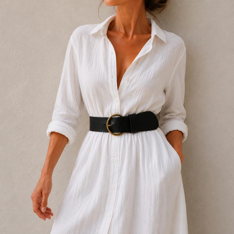 Puede incluir: Un vestido camisero blanco de manga larga con acabado de tela texturizada, que presenta un cintur&oacute;n negro ancho con una hebilla circular de tono lat&oacute;n ajustado a la cintura. El vestido tiene un cuello cl&aacute;sico y mangas remangadas.
