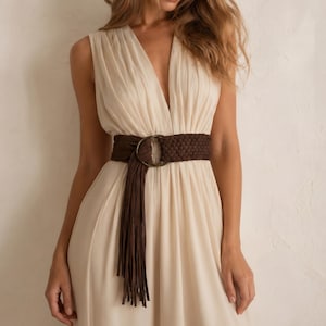 May include: A sleeveless, cream-colored dress with a deep V-neck is cinched at the waist with a wide, brown braided belt. The belt features a large, circular buckle and a long fringe detail that hangs down the front of the dress.