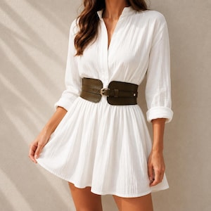 May include: A white long-sleeved mini dress with a textured fabric, featuring a V-neckline and a wide, dark olive green corset-style belt with a central buckle cinched at the waist. The dress has a flared skirt and cuffed sleeves.