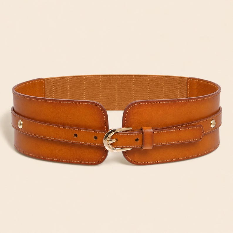 May include: A wide tan leather belt featuring a central gold-toned metal buckle and a matching leather strap overlay. The belt includes two small gold-toned metal studs on each side and an elasticated interior band for a flexible fit.