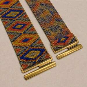 Puede incluir: Dos cinturones tejidos coloridos con patrones geom&eacute;tricos. Uno presenta un patr&oacute;n de diamantes repetido en azul, amarillo, naranja y gris. El otro tiene un patr&oacute;n mixto en naranja, azul y amarillo. Ambos cinturones tienen cierres met&aacute;licos dorados.