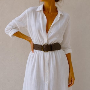 Puede incluir: Un vestido camisero blanco de manga larga con botones y tejido texturizado. El vestido se ajusta a la cintura con un cintur&oacute;n ancho de cuero marr&oacute;n oscuro que presenta una hebilla circular grande en tono lat&oacute;n y un cierre de hebij&oacute;n.