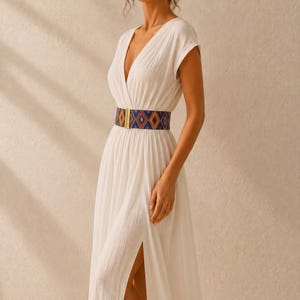 Puede incluir: Un vestido blanco con cuello en V y mangas cortas, con una abertura alta. La cintura se realza con un cintur&oacute;n con un estampado geom&eacute;trico en azul, naranja y dorado. La hebilla del cintur&oacute;n es dorada.