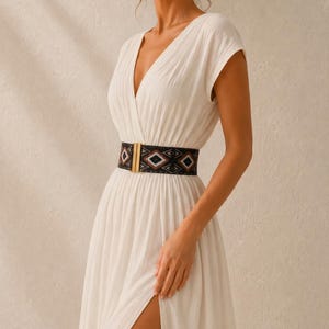 Puede incluir: Un vestido blanco con cuello en V, mangas cortas y una abertura alta. El vestido se ci&ntilde;e a la cintura con un cintur&oacute;n estampado en negro y marr&oacute;n con una hebilla dorada. El cintur&oacute;n presenta dise&ntilde;os geom&eacute;tricos.