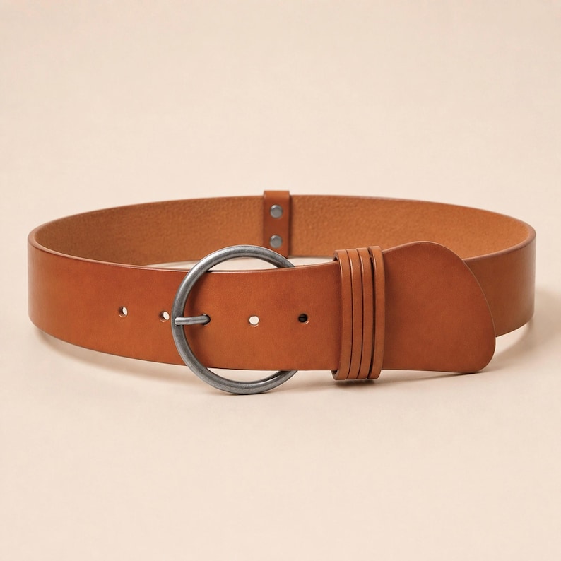 Puede incluir: Un cintur&oacute;n de cuero color canela con una hebilla circular grande de metal en tono plateado mate. La correa del cintur&oacute;n incluye tres trabillas decorativas verticales cerca de la hebilla y una serie de agujeros de ajuste.