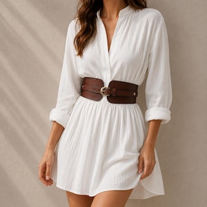 May include: A white long-sleeved dress with a textured fabric, featuring a V-neckline and a wide, dark brown leather-style corset belt with a gold-toned buckle cinched at the waist. The dress has a flared skirt and cuffed sleeves.