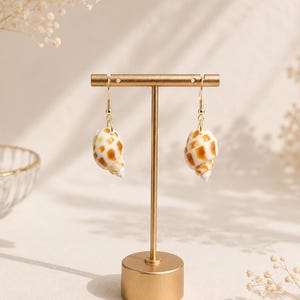 May include: A pair of seashell earrings with gold-colored hooks and a gold-colored display stand. The earrings are white with brown spots. The stand has a round base and a vertical post.