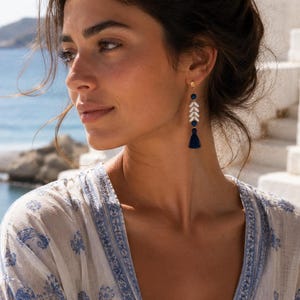 May include: A long drop earring featuring a gold-tone stud, a small dark blue bead, a vertical row of white leaf-shaped components, another dark blue bead, and a dark blue thread tassel at the bottom, hanging from a gold-tone metal hook.
