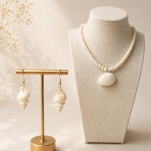 Könnte beinhalten: Ein Schmuckset mit Muschelmuster, bestehend aus einer Halskette und Ohrringen. Die Halskette hat ein Perlendesign mit einem Muschelanhänger. Die Ohrringe sind muschelförmig und hängen an goldfarbenen Haken. Der Schmuck wird auf einer cremefarbenen Büste und einem goldenen Ohrringständer präsentiert.