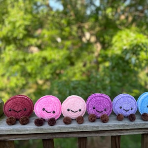 May include: A row of seven small, round, crocheted plush toys in a gradient of colours including dark red, pink, light pink, purple, lavender, blue, and teal. Each plush features a smiling face with black eyes and two small brown feet.