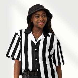 May include: A black and white striped short-sleeve shirt with a black collar and buttons. The shirt is paired with a black bucket hat. The shirt's design features bold, vertical stripes, creating a striking contrast.