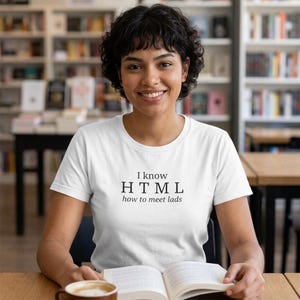 Könnte beinhalten: Ein weißes T-Shirt mit dem Text "I know HTML how to meet lads" in Schwarz. Die Person lächelt und sitzt an einem Tisch mit einem Buch und einer Kaffeetasse. Der Hintergrund umfasst Bücherregale.
