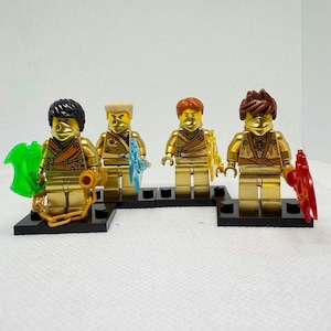 May include: Four gold-colored Lego minifigures with various hair colors and accessories. Each figure stands on a black base and holds a different weapon, including a green axe, a blue shield, and a red blade. The figures have gold masks.