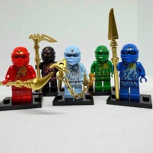 May include: A collection of six Lego Ninjago minifigures. Each figure is a different color: red, black, light blue, green, and dark blue. They are all wearing ninja masks and carrying golden weapons. The figures are standing on black baseplates.