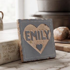 Op de afbeelding: Een vierkante grijze stenen plaat met een uitgesneden hartvorm in het midden. Binnenin het hart is de naam EMILY gegraveerd in dikke grijze letters, met een kleiner, effen grijs hart direct onder de naam op een lichtbruine achtergrond.