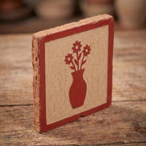Puede incluir: Un azulejo cuadrado de terracota con un borde rojo en relieve y un diseño central grabado de un jarrón con tres flores. El azulejo tiene una superficie texturizada de color beige claro con acabado mate y un borde rugoso de arcilla.