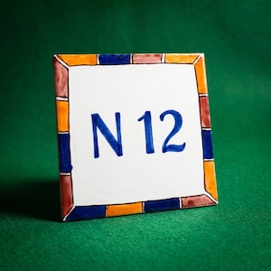 May include: A square ceramic tile featuring the blue painted text N 12 in the centre. The tile has a white background and a decorative border composed of alternating rectangular segments in shades of orange, dark blue, and muted brown.