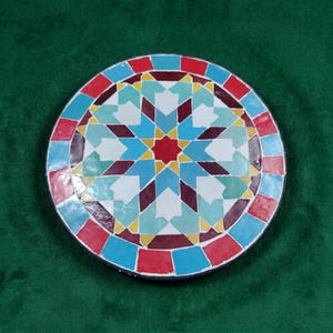 Könnte beinhalten: Ein kreisförmiges Mosaikstück mit einem geometrischen Sternmuster in der Mitte. Das Design umfasst eine lebendige Farbpalette aus roten, weißen, gelben, hellblauen und dunkelroten Fliesen, die in einem symmetrischen Sternmotiv angeordnet sind.
