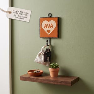 May include: A square wall-mounted hook featuring an orange tile with a white heart graphic and the name AVA printed in the centre. Below the hook, a small wooden shelf holds a terracotta-coloured bowl with keys and a small potted succulent plant.