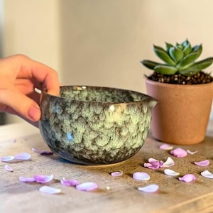 May include: A ceramic bowl with a textured, mottled green and black glaze. The bowl has a small spout and is held by a hand. A small potted succulent plant sits nearby. Scattered purple flower petals are on the wooden surface.
