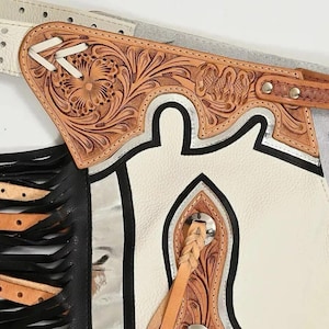 Op de afbeelding: Close-up van een witte en zwarte leren chaps met ingewikkelde bloemen- en scrollbewerking. De chaps hebben zilveren accenten, een decoratieve leren riem en zwarte franjes. Het leer is lichtbruin van kleur.