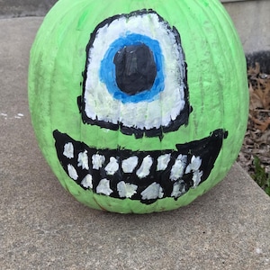 May include: A green pumpkin painted with a large, cartoonish eye with blue and black details. The pumpkin also features a wide, toothy grin. The image is taken on October 31, 2025, at 5:11 PM in Warrenton.
