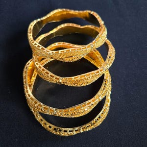 May include: Four gold bangles with detailed filigree and textured patterns are stacked on a dark blue surface. The bangles have a bright, polished finish, reflecting light. The overall aesthetic is ornate and elegant, suitable for special occasions.