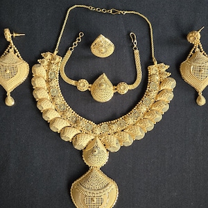 Op de afbeelding: Een complete goudkleurige sieradenset, inclusief een ketting, oorbellen, ring en armband, allemaal met ingewikkelde filigraan- en textuurontwerpen. De ketting heeft een grote hanger en een bijpassende armband. De oorbellen zijn druppelvormig.