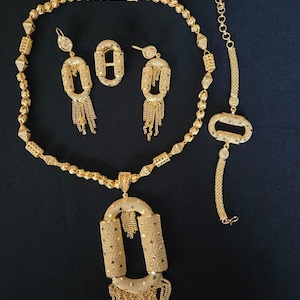Könnte beinhalten: Ein goldfarbenes Schmuckset mit Halskette, Ohrringen, einem Ring und einem Armband. Die Halskette hat einen großen Anhänger mit rechteckigem Design und baumelnden Ketten. Die Ohrringe und das Armband haben ebenfalls rechteckige Designs. Das Schmuckstück liegt auf schwarzem Grund.