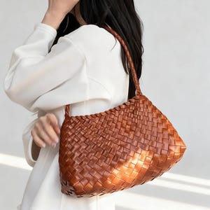 Puede incluir: Un bolso de hombro de cuero trenzado marrón con asa trenzada. El bolso tiene forma rectangular y un patrón tejido. El bolso es de tamaño mediano y lo sostiene una persona.