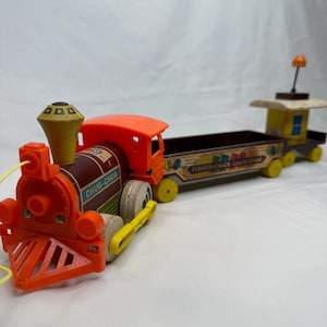 May include: Vintage Fisher-Price Chug-Chug train toy. The toy train is primarily orange, brown, and yellow. The train has a red cab and a brown car with the words "Fisher-Price Chug-Chug" printed on the side. The train is made of plastic and wood.