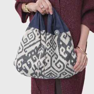 May include: A patterned tote bag with a navy blue top and a decorative button. The bag features a cream and grey geometric design. The bag is held by a person wearing a burgundy cardigan and gold bracelets.