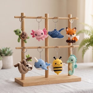 May include: A wooden display stand holding eight crocheted animal keychains. The collection includes a green turtle, pink axolotl, blue stingray, orange fox, brown sloth on a branch, blue narwhal, yellow and black bee, and a light blue seahorse.