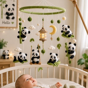 May include: A nursery mobile featuring crocheted panda bears, green bamboo stalks, white clouds, a yellow crescent moon, and a central beige lantern. The items hang from a circular green frame suspended by white strings from a wooden arm.