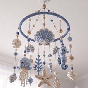 May include: A hanging nursery mobile featuring crocheted sea creatures including a jellyfish, seahorse, starfish, and various seashells in shades of blue, cream, and beige. The items are suspended from a blue circular frame with small wooden beads.