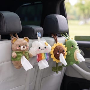 May include: Four crocheted tissue holders hanging from car headrests. The set includes a brown bear in a green vest, a white cloud with a rainbow, a yellow sunflower, and a green dinosaur. Each figure features a central opening for tissues.
