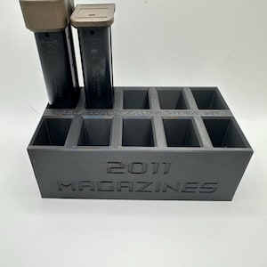 Può includere: Un porta caricatore nero 2011 con due caricatori. Il supporto ha fessure per più caricatori e il testo "2011 MAGAZINES" è impresso sul davanti. I caricatori sono neri con la parte superiore color cuoio.