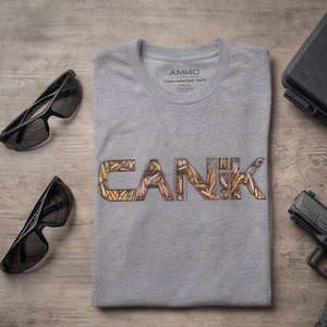 May include: A gray t-shirt with the word "CANIK" in a bullet-filled design. The shirt has a crew neck and the brand name "AMMO" is printed on the collar. Black sunglasses and a handgun are visible.