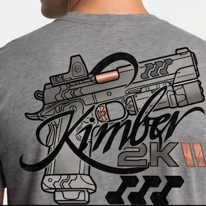 Puede incluir: Camiseta gris con un gráfico detallado de una pistola y el texto "Kimber 2K III". El diseño incluye una mira de punto rojo y detalles en color cobre. La camiseta es de color gris liso.