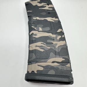 Puede incluir: Un cargador de rifle de polímero con un patrón de camuflaje negro y beige. El cargador tiene una superficie de rejilla texturizada y el texto en relieve PMAG 30 cerca de la base. Es un componente curvo y rectangular para almacenar munición.