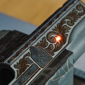 Może przedstawiać: Metalowy zamek broni palnej z misternymi grawerunkami w formie zwojów oraz logo Springfield Armory 1794. Na powierzchni metalu widoczna jest mała, jasnopomarańczowa kropka lasera, podkreślająca szczegółowe wykonanie tego wzoru.