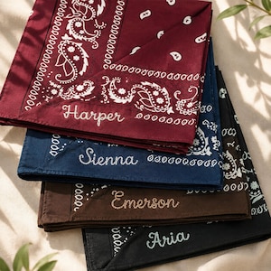 May include: A stack of four folded bandanas in burgundy, navy blue, brown, and black. Each bandana features a white paisley pattern and is embroidered with a name in cursive script: Harper, Sienna, Emerson, and Aria, respectively.