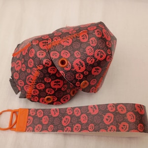 Può includere: Un portachiavi da polso e una pochette arrotondata abbinata in similpelle nera e rossa con un motivo ripetuto di zucche. La pochette presenta cuciture e occhielli metallici arancioni, mentre il portachiavi include un anello in plastica arancione.