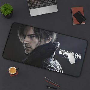 May include: A rectangular black mousepad featuring a portrait of a man with dark hair and the text "RESIDENT EVIL" in white. A white computer mouse rests on the pad. A laptop, coffee cup, and pencils are also visible.
